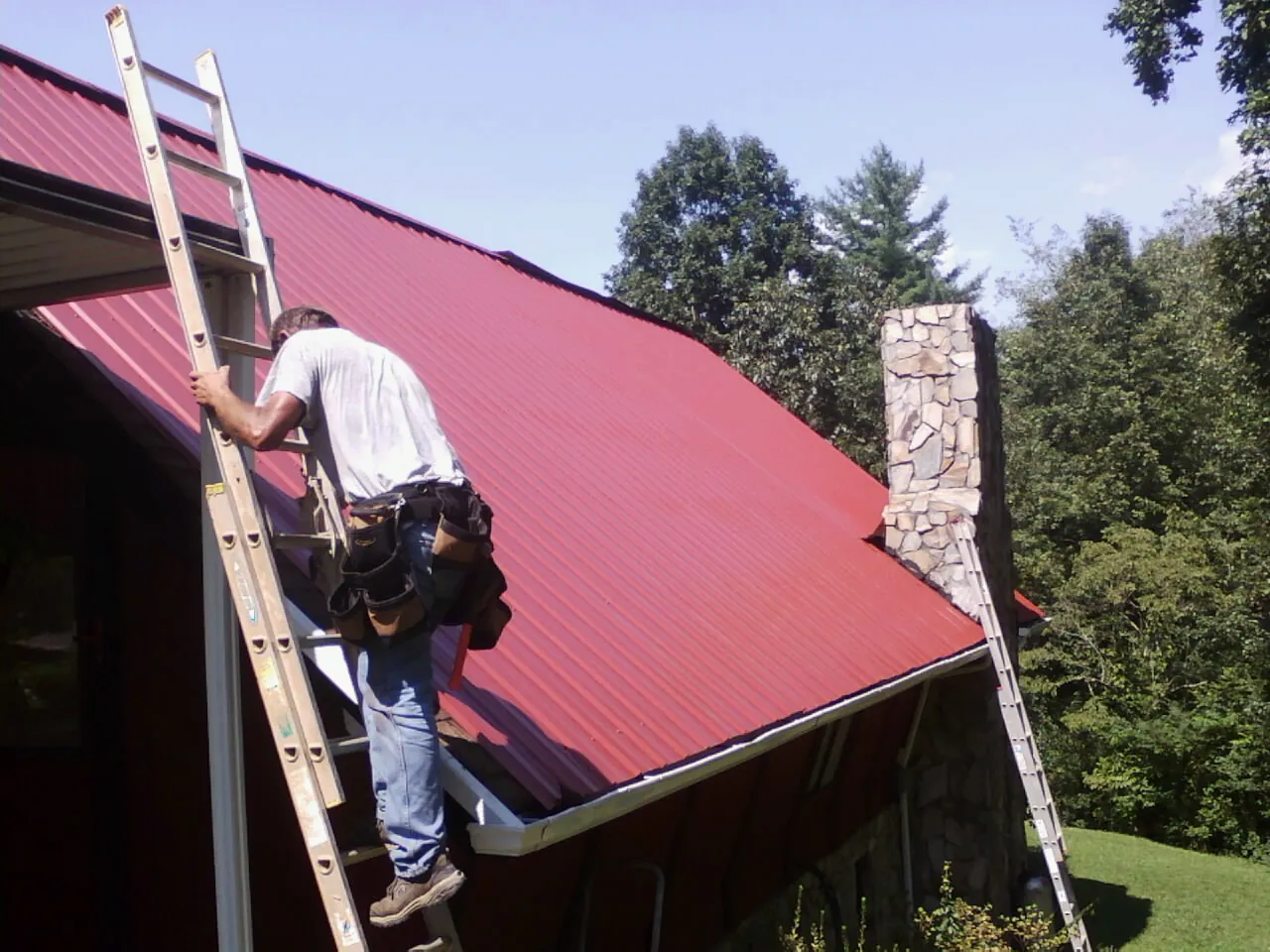 Roofer on a ladder beside a red metal roof and stone chimney — Soffit & Fascia Repair by Roofing Brothers in Barry